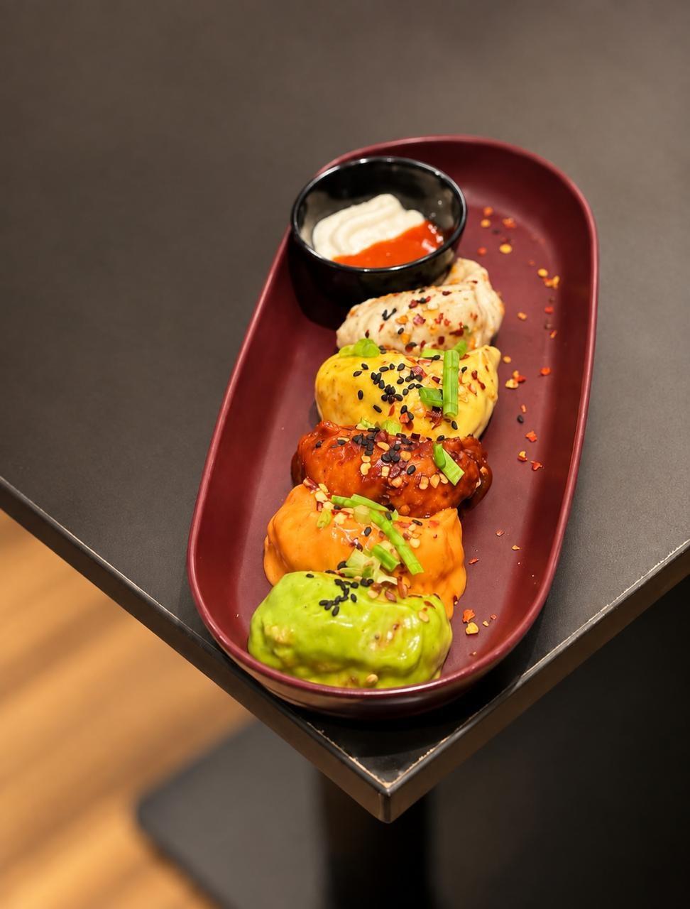 A red platter of five momos in different coloured sauces with chilli flakes, sesame and a dip