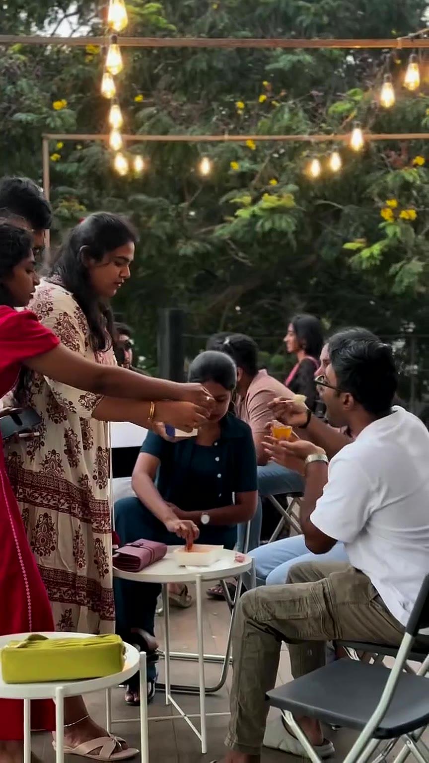 Friends sharing food around small tables under string lights at dusk