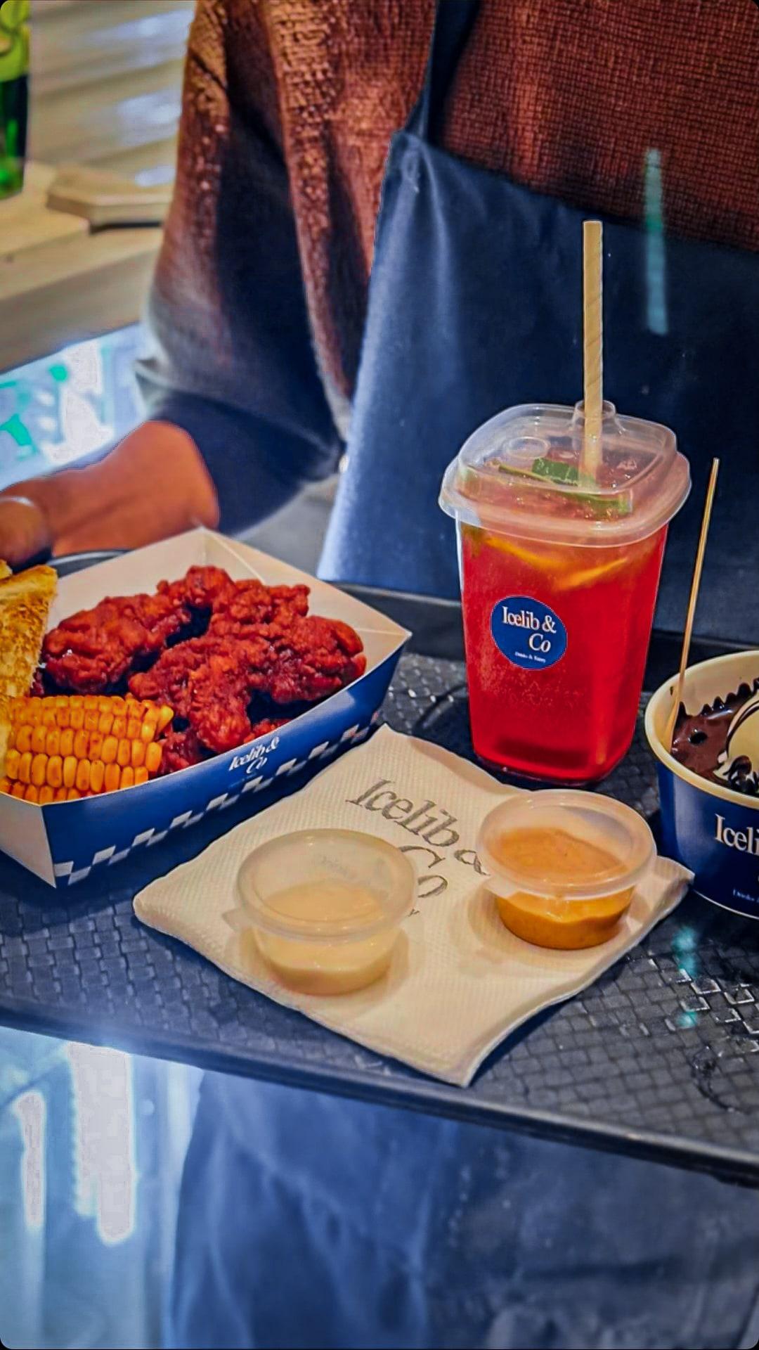 A server holding a tray of Nashville chicken tenders with corn and toast, a red iced tea, dipping sauces and a mud bowl, all in Icelib & Co packaging